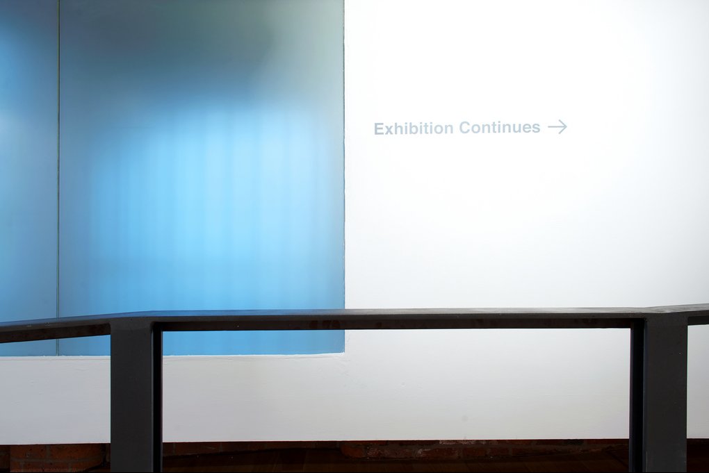 Exhibition Continues signage — translucent blue glass panel, steel railing